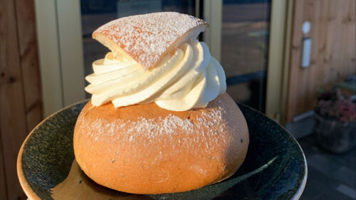 En hand håller i en tallrik med en semla på, framför en dörr med texten Leijon Bageri
